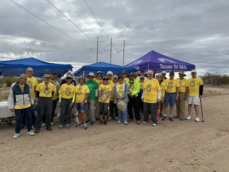 About twenty volunteers in matching yellow shirts standing together under canopies at a desert aid station.