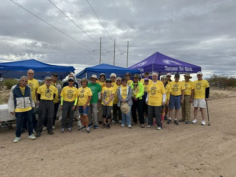 About twenty volunteers in matching yellow shirts standing together under canopies at a desert aid station.