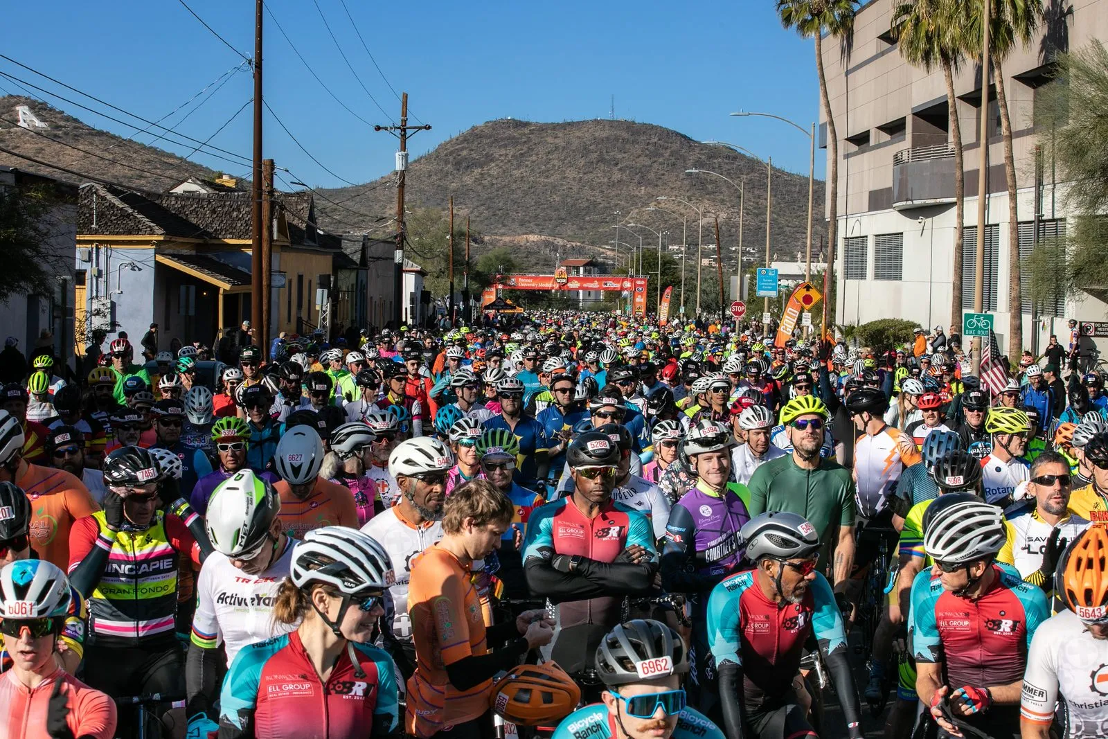 Riders packed shoulder to shoulder filling a Tucson street to the horizon, adobe row houses on one side and a mountain behind.