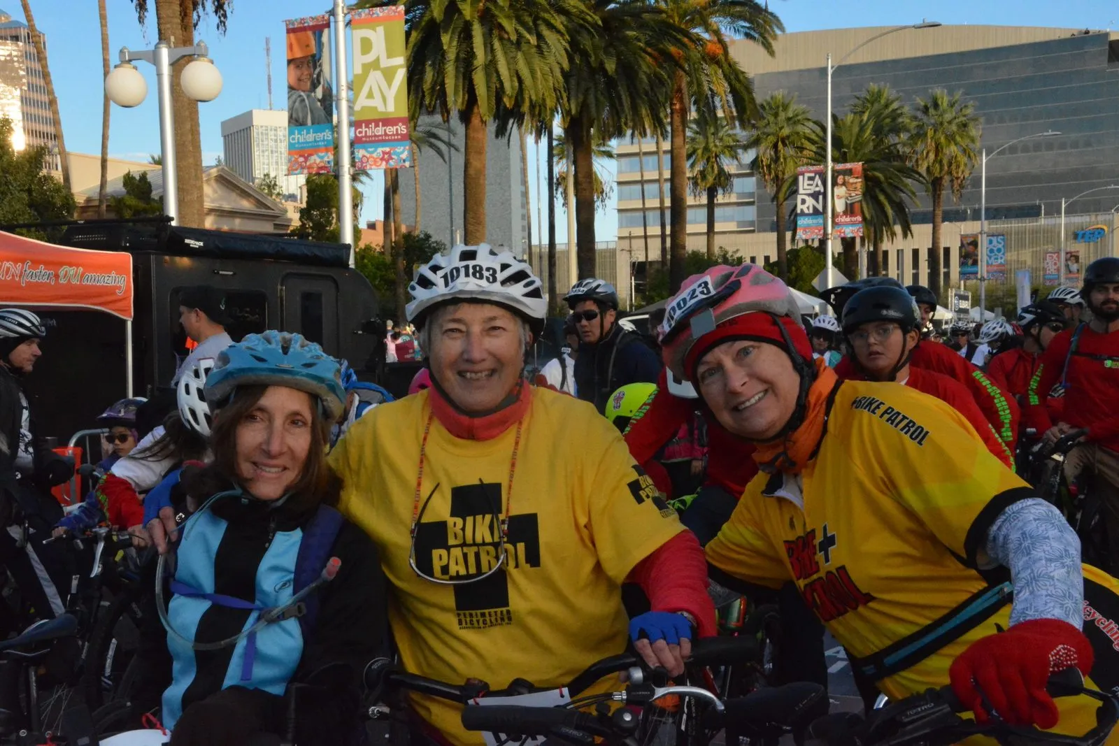 Three Bike Patrol volunteers with their arms around each other, two in yellow patrol jerseys and one in a blue jacket, riders packed into the street behind them.