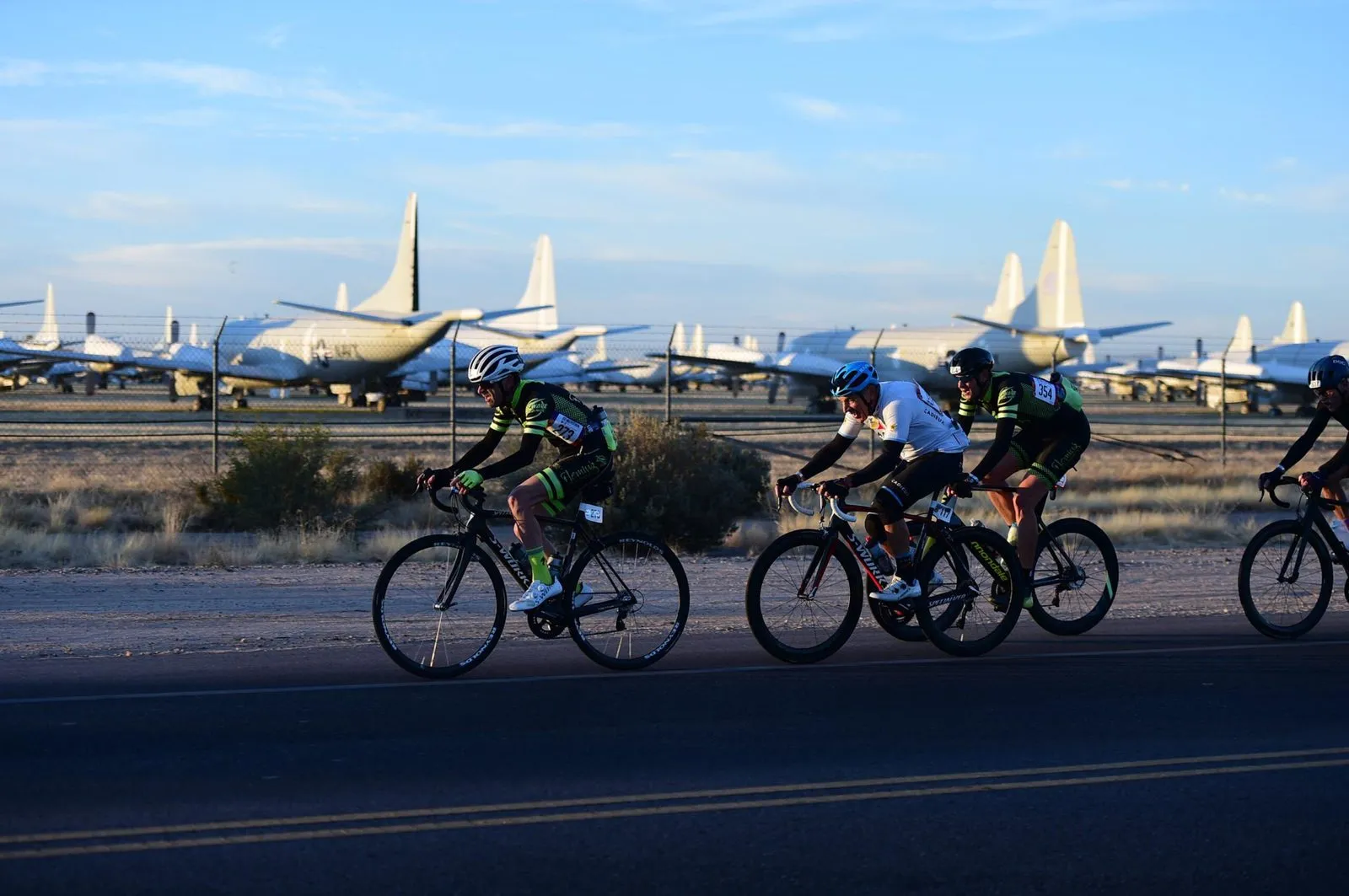 A line of riders on a highway with rows of mothballed military aircraft filling the background.