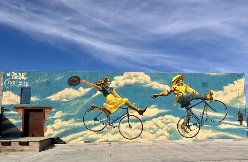 A mural on a downtown Tucson wall showing two riders on antique bicycles floating through clouds.