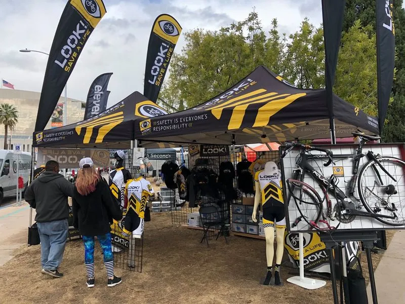 A bike brand booth with a road bike on a display grid, apparel racks and customers looking on.