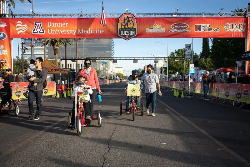 Two children on adaptive trikes rolling under the finish arch with families walking alongside.