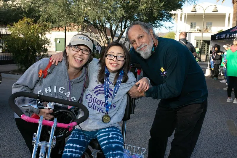 A girl on an adaptive handcycle wearing a finisher medal, hugged by two smiling adults.