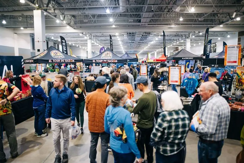 A crowd browsing merchandise tables under El Tour canopies inside a convention center hall.