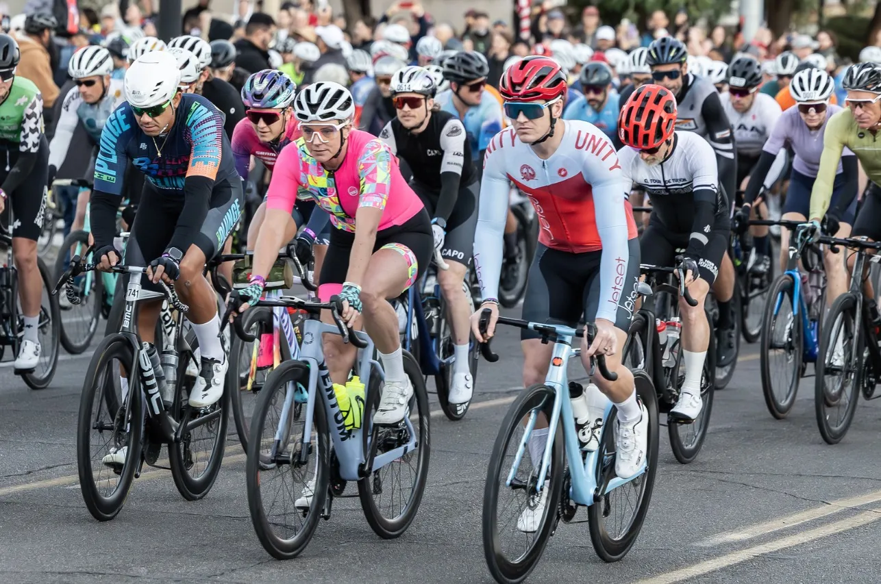 A pack of riders rolling down a city street toward the camera, a rider in a bright pink jersey at the front, spectators along the far curb.