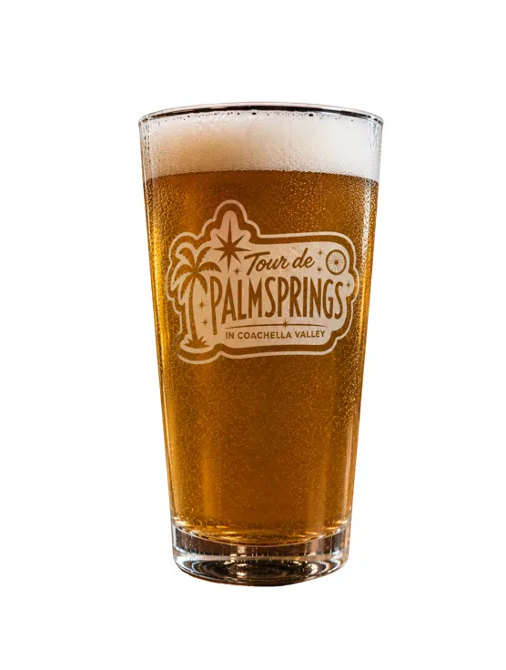 Pint glass of beer with the Tour de Palm Springs logo printed on the front, on white