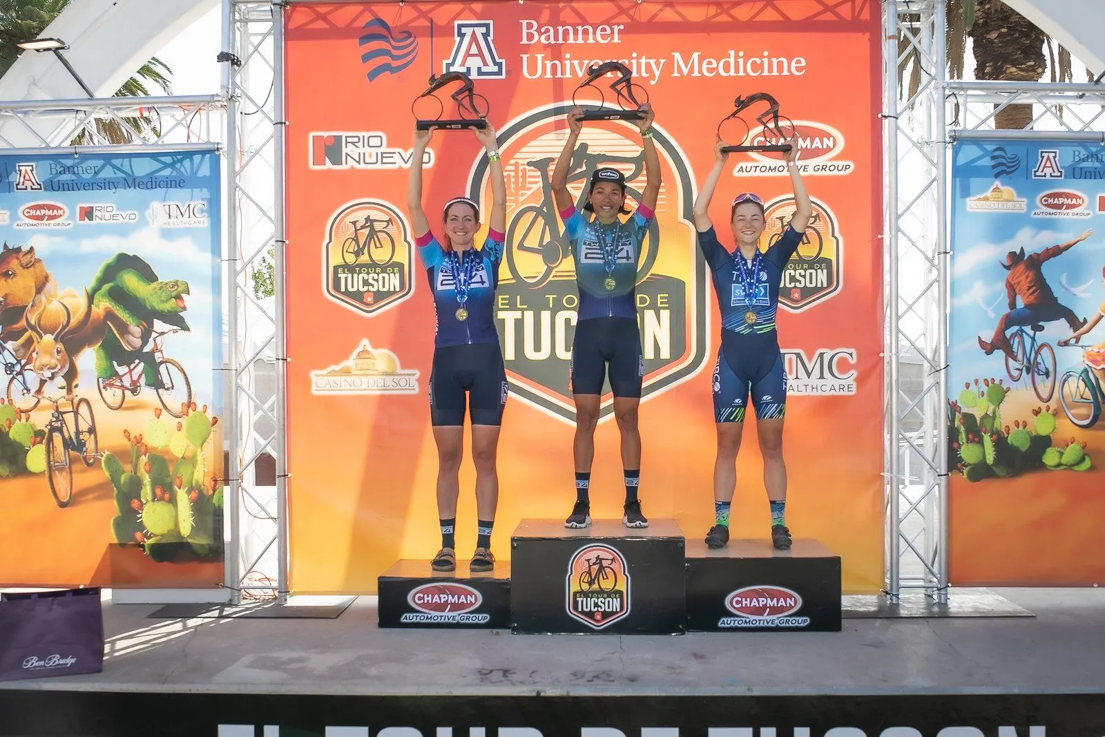 Three women on a podium raising bicycle-shaped trophies overhead.