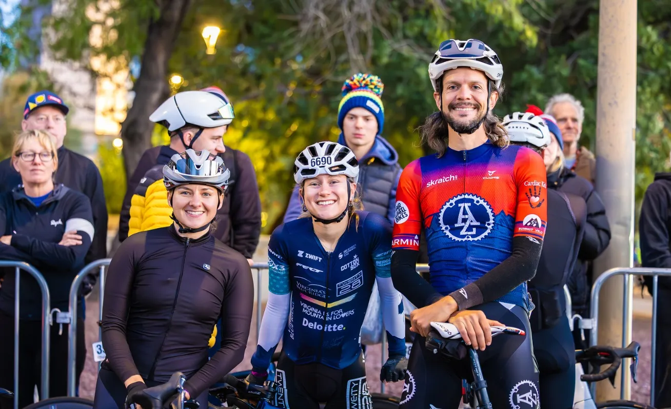 Three riders standing over their bikes in early morning light, smiling at the camera, with spectators behind the barriers.
