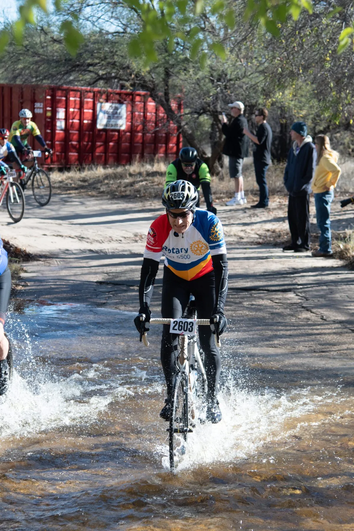 A rider riding through a flooded road, water spraying from the wheels, spectators watching from the bank.