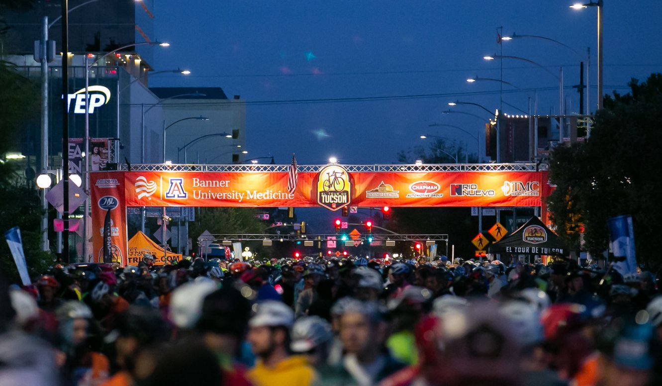 Cyclists at a mass start rolling under a lit start gantry at blue hour.